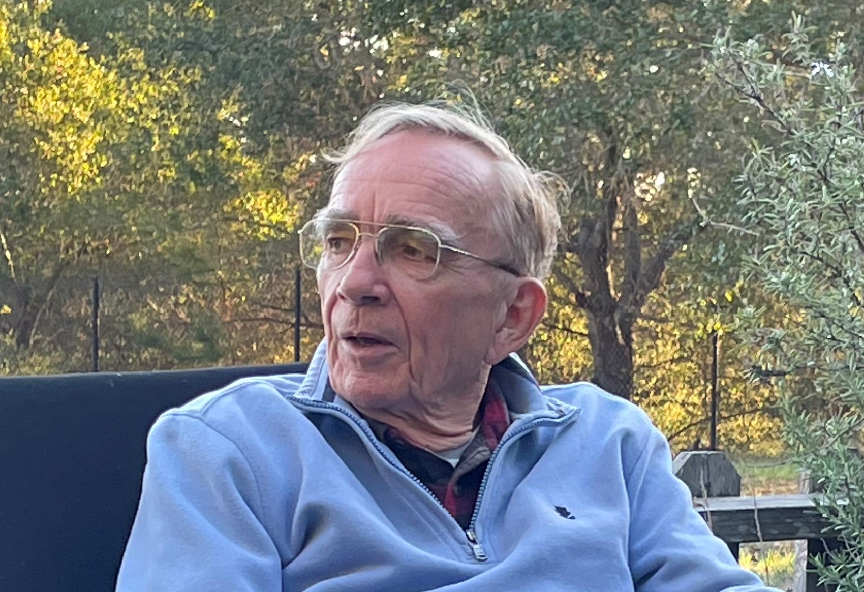 Dr. Eaton in a blue sweatshirt and gold glasses sitting. He is engaged in conversation and trees fill the background.